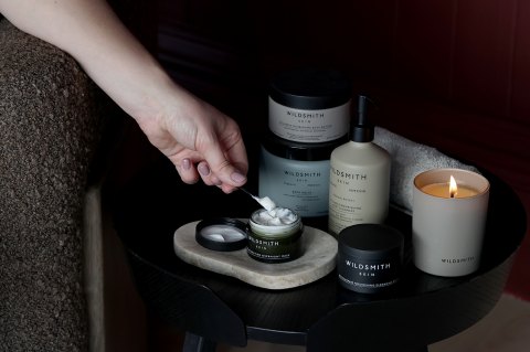 Luxury Wildsmith lit candle and skin care products on a black bedside table, a ladies hand scooping the contents of face cream